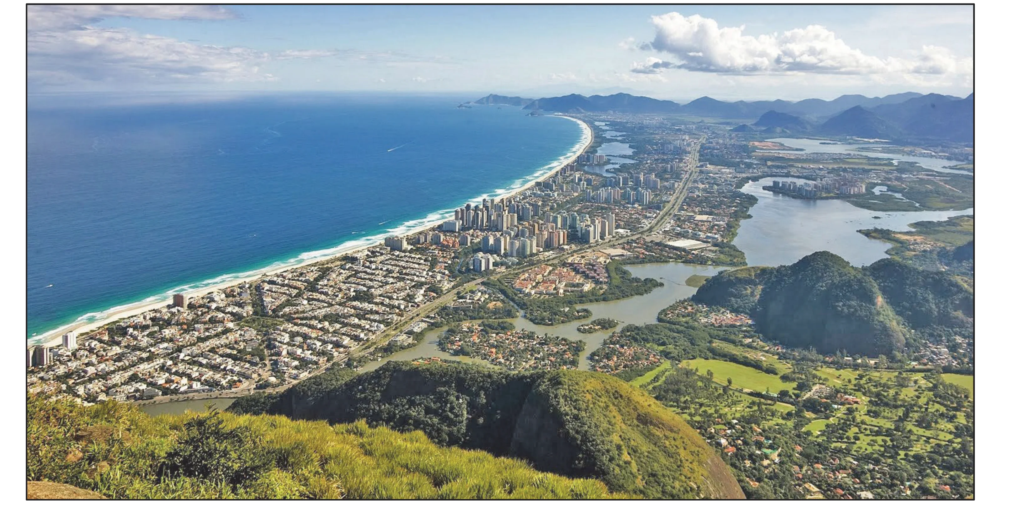 Widok z góry na fragment wybrzeża Rio de Janeiro (23 stopnie S, 43 stopnie W), obejmujący długą piaszczystą plażę, ocean z falami, gęstą miejską zabudowę, jeziora i zielone góry w tle.